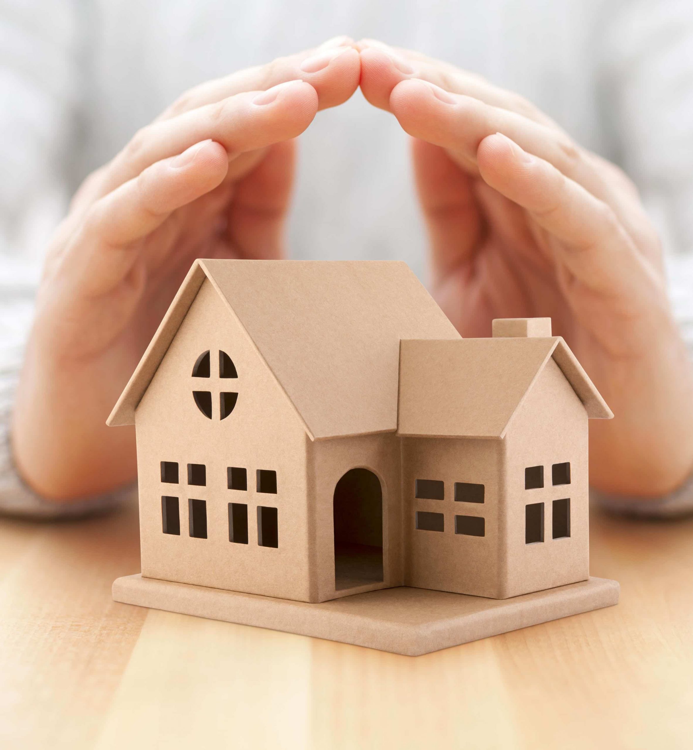 Modèle miniature d'une maison protégée par des mains placées en forme d'arc, représentant la protection et la sécurité d'une assurance habitation