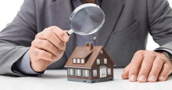 contre expertise en assurance habitation Un homme en costume examine une petite maquette de maison à l'aide d'une loupe, symbolisant le processus de contre-expertise en assurance habitation.